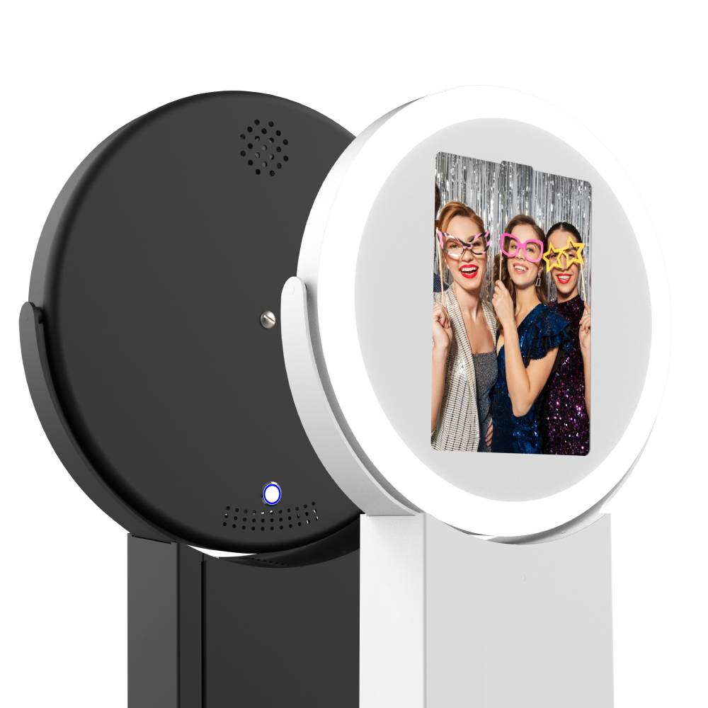 A front and rear view of two modern circular photo booth kiosks; the white unit displays three women posing with props on its screen, while the matte black unit shows a rear speaker grille and a glowing blue power button.