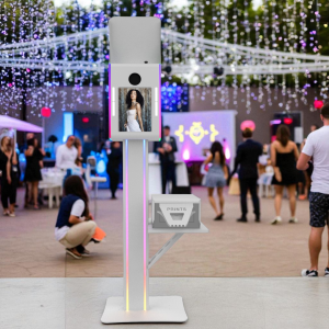 A white M1 photo booth standing in a decorated outdoor event space, featuring an integrated vertical monitor, top flash bounce card, and a printer shelf with a "Prints" cover.