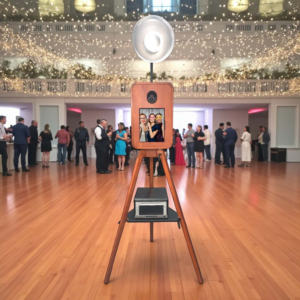 A wooden, vintage-style photo booth on a tripod stand with a large silver flash reflector, set in a ballroom.
