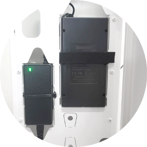 A close-up view inside a white photo booth housing, showing a black power adapter with a green indicator light and a large "TalentCell" lithium-ion battery pack secured with a black Velcro strap.