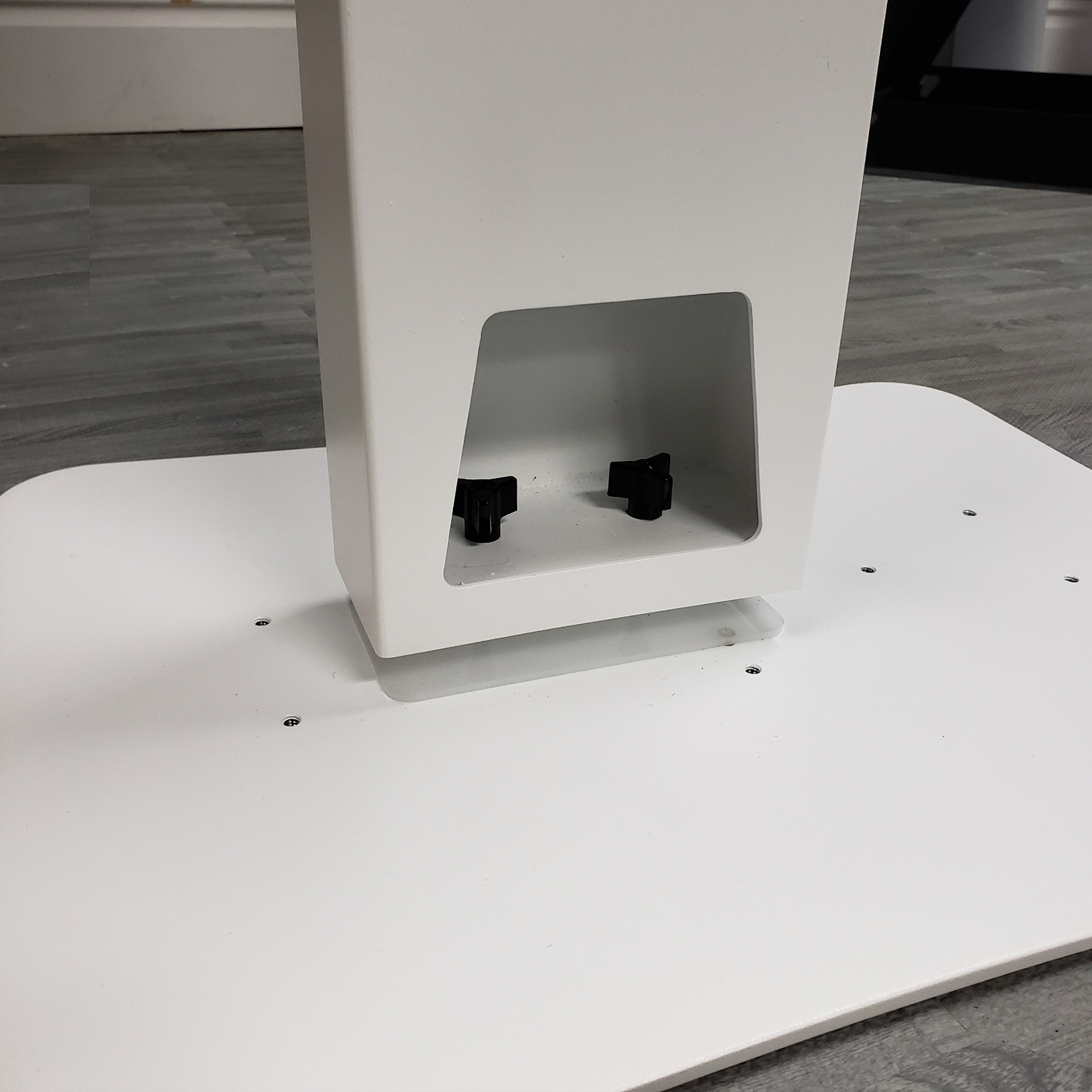 A close-up view of the base of a white photo booth column, showing two black wing nuts used for tool-less assembly to the flat base plate.